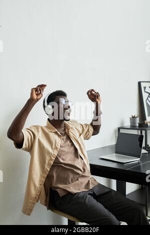Homme d'affaires africain heureux dans des casques sans fil dansant tout en étant assis à son lieu de travail avec un ordinateur portable au bureau Banque D'Images