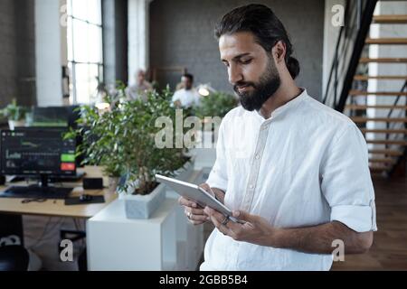 Jeune homme d'affaires arabe en chemise blanche regardant l'écran de tablette tout en faisant défiler les données en ligne Banque D'Images