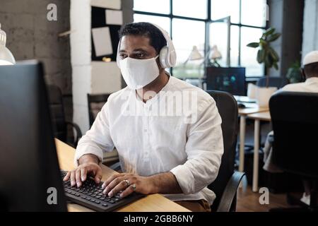 Homme d'affaires sérieux de course mixte dans le masque et le casque regardant l'écran d'ordinateur tout en tapant Banque D'Images
