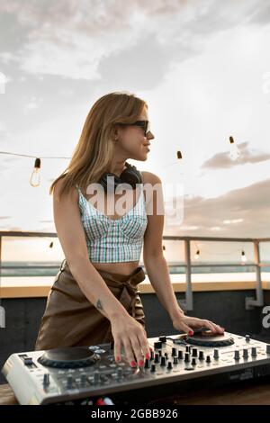 Bonne jeune femme blonde dans des lunettes de soleil debout près d'une table de son sur le patio sur le toit Banque D'Images