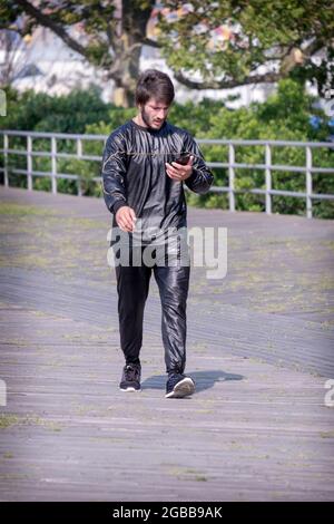 Lors d'une journée d'été très chaude à New York, un jeune homme fait de l'exercice en costume de survêtement et lit son téléphone cellulaire. À Brighton Beach, Brooklyn, New york. Banque D'Images