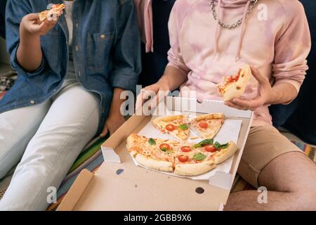 Deux adolescents décontractés en vêtements décontractés qui mangent de la pizza de la boîte tout en étant assis sur le sol Banque D'Images