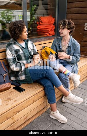 Un couple adolescent en vêtements décontractés assis sur le banc près d'un bâtiment moderne, parlant et prenant des boissons Banque D'Images