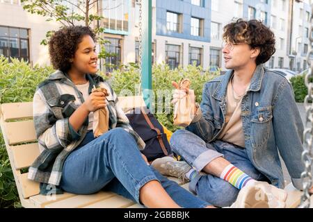 Jeune couple interculturel en vêtements décontractés prendre un verre tout en se relaxant sur les balançoires à l'extérieur Banque D'Images