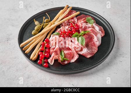 Gros plan sur le prosciutto coppa di parma tranches de jambon avec basilic, bâtonnets de grissini, câpres et groseilles sur plaque sombre sur fond texturé Banque D'Images