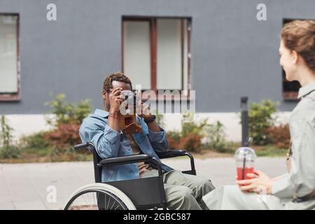 Photographe afro-américain assis en fauteuil roulant et utilisant un appareil photo traditionnel tout en prenant la photo d'une femme avec un verre Banque D'Images