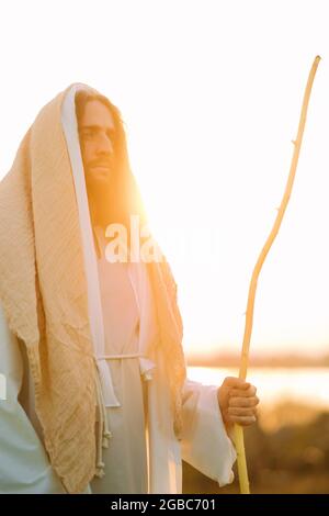 Jésus Christ avec un personnel en bois se tient dans un pré vêtu de sa robe blanche traditionnelle contre fond de rivière et de coucher de soleil. Banque D'Images