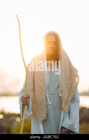 Jésus Christ avec un personnel en bois se tient dans un pré vêtu de sa robe blanche traditionnelle contre fond de rivière et de coucher de soleil. Banque D'Images