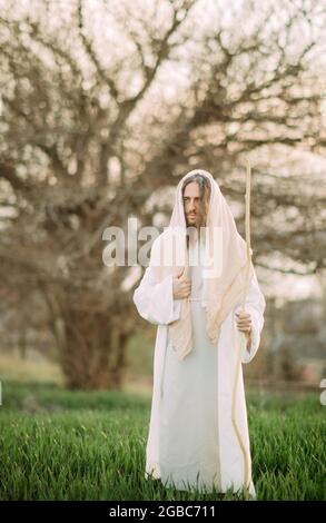 Jésus Christ avec un personnel en bois se tient dans un pré vêtu de sa robe blanche traditionnelle sur fond d'arbre. Banque D'Images