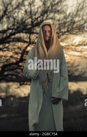 Jésus Christ marche dans la prairie vêtu de sa robe blanche traditionnelle contre le ciel et le fond de l'arbre au coucher du soleil. Banque D'Images