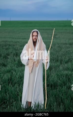 Jésus Christ avec le personnel en bois marche dans la prairie vêtu de sa robe blanche traditionnelle. Banque D'Images