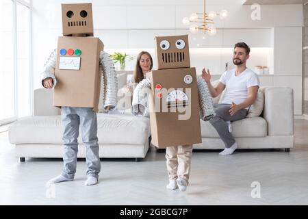 Famille heureuse de parents et de deux enfants jouant à la danse comme des robots à la maison, les enfants portant un costume de boîte mobile fait main en carton Banque D'Images