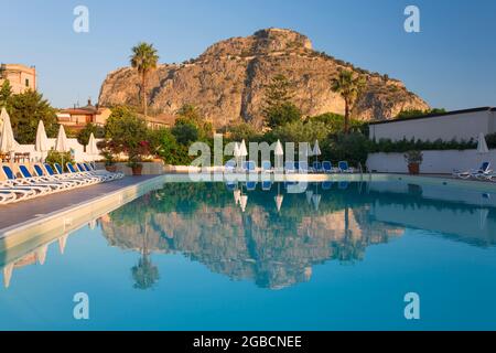 Cefalù, Palerme, Sicile, Italie. Les imposantes falaises ensoleillées de la Rocca se reflètent dans la piscine tranquille de l'hôtel Kalura, lever du soleil. Banque D'Images