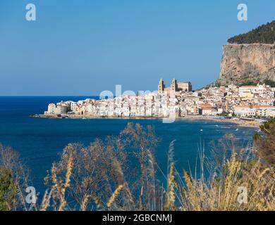 Cefalù, Palerme, Sicile, Italie. Vue sur la baie depuis la colline herbeuse, la ville et la cathédrale arabo-normande naine par les imposantes falaises de la Rocca. Banque D'Images