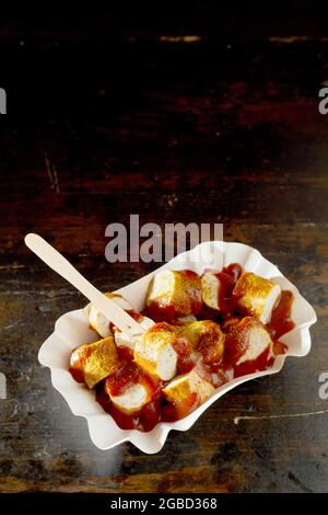 De part en part de savoureux morceaux de saucisse de porc frits avec ketchup au curry et bâton biologique dans un bol jetable sur fond sombre Banque D'Images