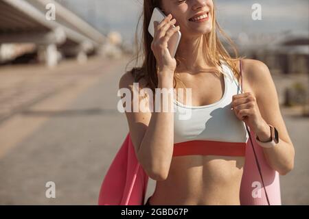 Une jeune femme mince en costume de piste sourit en parlant sur un téléphone portable dans la rue de près Banque D'Images