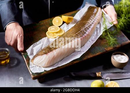 Crop anonyme chef en uniforme noir démontrant filet de morue non cuit et tranches de citron disposées sur un panneau en bois dans la cuisine Banque D'Images