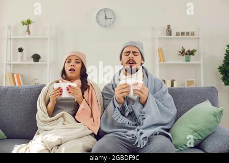Jeune couple dans des vêtements chauds et des couvertures assis sur un canapé avec des mouchoirs, prêt à éternuer et se sentir mal Banque D'Images