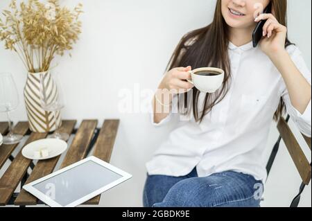 Une femme dans une chemise blanche et un Jean est assise dans un café et boit du café. Une employée de bureau à la pause déjeuner parlant au téléphone. Responsable à un Banque D'Images