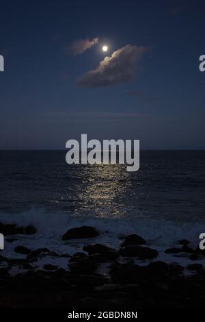 Une pleine lune brille sur une nuit claire au-dessus de la mer et de la côte rocheuse dans ce paysage de nature. Banque D'Images