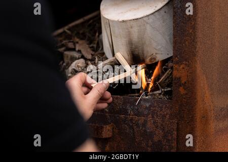 Homme à la main faisant un feu avec de petites branches en bois et une ancienne boîte en bois dans un poêle en métal rouillé extérieur tout en se relaxant et en appréciant la vie simple Banque D'Images