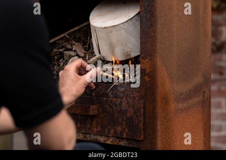 Un homme faisant un feu avec de petites branches en bois et une ancienne boîte dans un poêle en métal rouillé extérieur tout en se relaxant et en appréciant la vie simple Banque D'Images