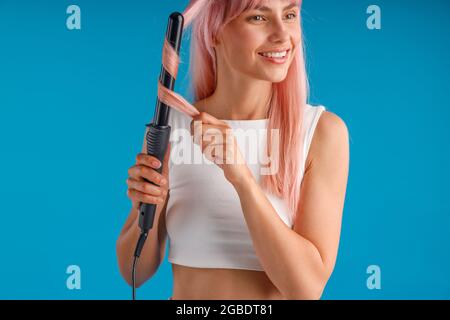 Belle femme souriante coiffant les cheveux longs roses à l'aide d'un fer à friser, debout isolé sur fond bleu studio Banque D'Images