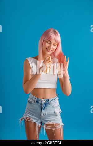 Jeune femme gaie souriant et appliquant un spray hydratant à ses beaux cheveux teints rose lisse tout en se tenant isolé sur le studio bleu Banque D'Images