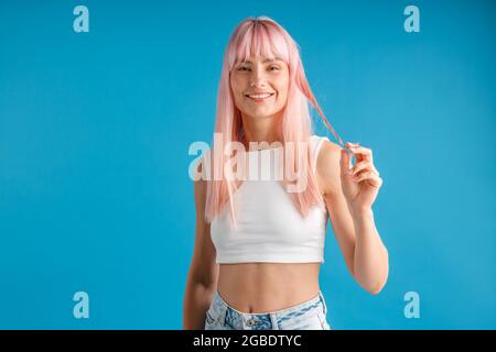 Jeune femme souriante avec de longs cheveux teints roses naturels tenant une mèche de cheveux et regardant la caméra, posant isolé sur fond bleu studio Banque D'Images