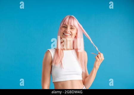 Jeune femme heureuse avec de longs cheveux teints roses naturels tenant une mèche de lui et souriant à la caméra, posant isolé sur fond bleu studio Banque D'Images