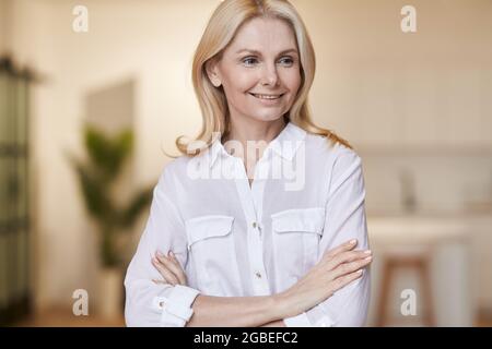 Femme réussie. Portrait d'une élégante femme mûre portant une chemise blanche en souriant de côté tout en posant avec les bras croisés pour l'appareil photo, debout à la maison Banque D'Images