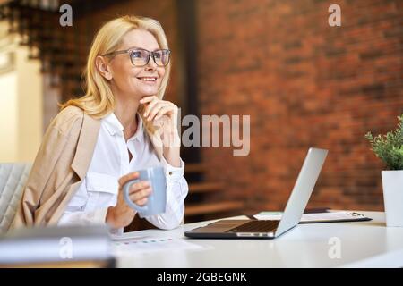 Femme d'affaires gaie et mûre dans des verres souriant, utilisant un ordinateur portable et buvant du café tout en travaillant au bureau Banque D'Images