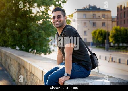 Jeune homme arabe indien ou hispanique en chemise noire avec un sac à bandoulière et des lunettes de soleil assis sur un mur en souriant tout en regardant l'appareil photo contre bl Banque D'Images