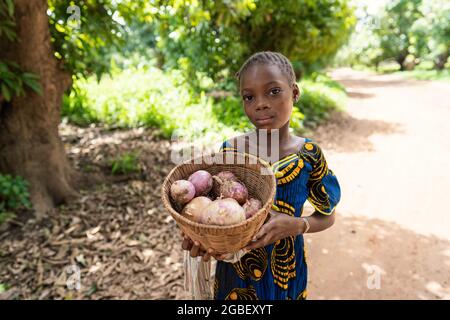 Dans cette image, une petite fille africaine noire sérieuse en robe traditionnelle se promette au marché du village avec un panier lourd rempli de gros oignons rouges Banque D'Images