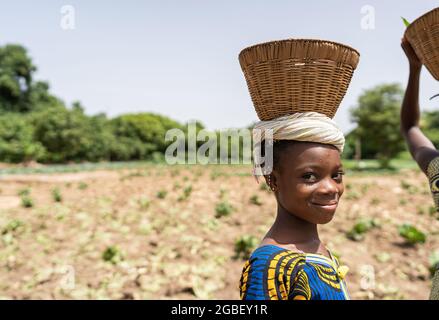 Gros plan d'une petite fille africaine souriante avec un panier vide sur sa tête revenant du marché du village après avoir vendu tous ses légumes Banque D'Images