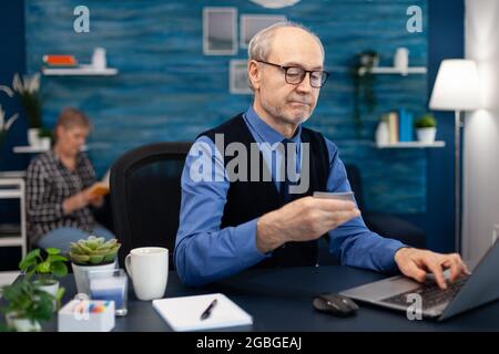 Homme senior utilisant une carte de crédit pour vérifier le compte bancaire. Homme âgé vérifiant les services bancaires en ligne pour faire un paiement en cheppping regardant l'ordinateur portable pendant que la femme est en train de lire un livre assis sur un canapé. Banque D'Images