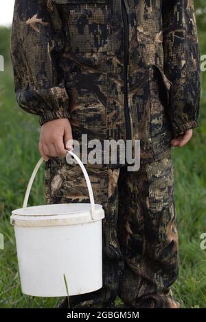 Gros plan du seau en plastique blanc dans les mains de l'enfant en costume de camouflage sur fond vert naturel. Voyage, vacances concept d'aventure. Banque D'Images