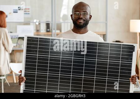 Portrait d'un ingénieur africain tenant une batterie solaire dans ses mains et regardant l'appareil photo tout en se tenant au bureau Banque D'Images