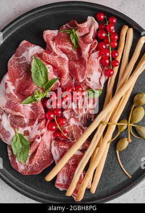 Fond alimentaire prosciutto coppa di parme jambon avec basilic, bâtonnets de grissini, câpres et groseille sur une assiette sombre. Orientation verticale Banque D'Images