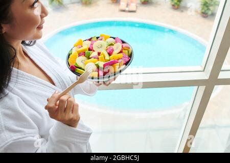 Positive jeune femme asiatique se tenant à la fenêtre panoramique avec vue sur la piscine et manger de la salade de fruits tropicaux Banque D'Images