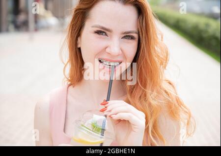 Jeune belle femme aux cheveux rouges avec bretelles boissons rafraîchissantes cocktail à l'extérieur en été. Portrait d'une fille souriante avec des taches de rousseur Banque D'Images