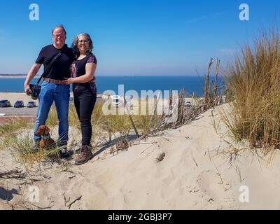 Les touristes seniors avec leur cachshund brun aux cheveux courts debout sur une colline de sable avec un parking avec des voitures et la mer du Nord en arrière-plan, Bro Banque D'Images