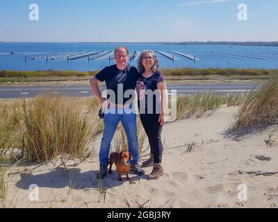 Couple multiculturel avec leur cachshund brun à poil court debout sur une colline avec une route et le lac Grevelingenmeer en arrière-plan, Brouwersda Banque D'Images