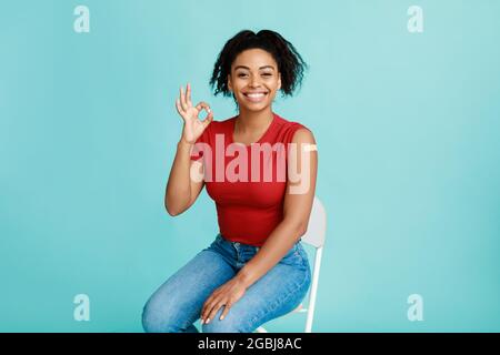 Joyeuse jeune femme noire avec bras vacciné après injection de covid montrant un geste correct et souriant à l'appareil photo Banque D'Images