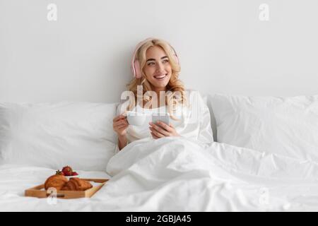 La jeune femme mange le petit déjeuner au lit écoute de la musique, aime le temps libre, les week-ends et les vacances Banque D'Images