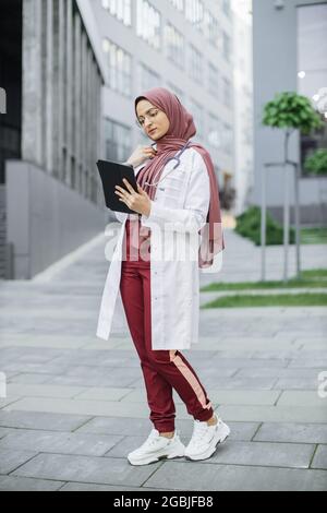 Femme musulmane médecin en hijab, portant des exfoliants et un manteau blanc, travaillant avec les dossiers des patients sur une tablette numérique en dehors de la clinique moderne Banque D'Images