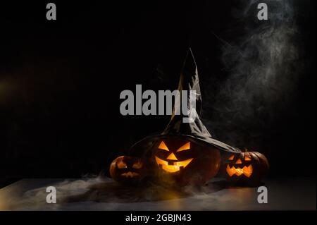 Les citrouilles d'orange effrayantes pour Halloween sont rangées sur un fond sombre. Jack O Lantern dans un chapeau de sorcière. Le brouillard mystique avance sur le sol. Trick or Banque D'Images