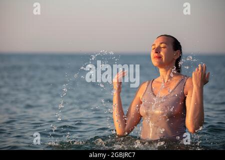 Une femme d'âge moyen se tient dans la mer et éclabousse l'eau. Banque D'Images