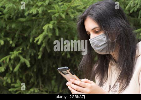 La fille dans le masque médical regarde le téléphone. Une femme aux longs cheveux noirs tient un téléphone portable. Soins de santé, covid19. Le port de masques dans publ Banque D'Images