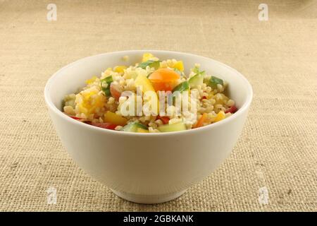 Salade de Bulgur fraîchement préparée avec fruits et légumes colorés dans un bol blanc sur fond hessien brun Banque D'Images